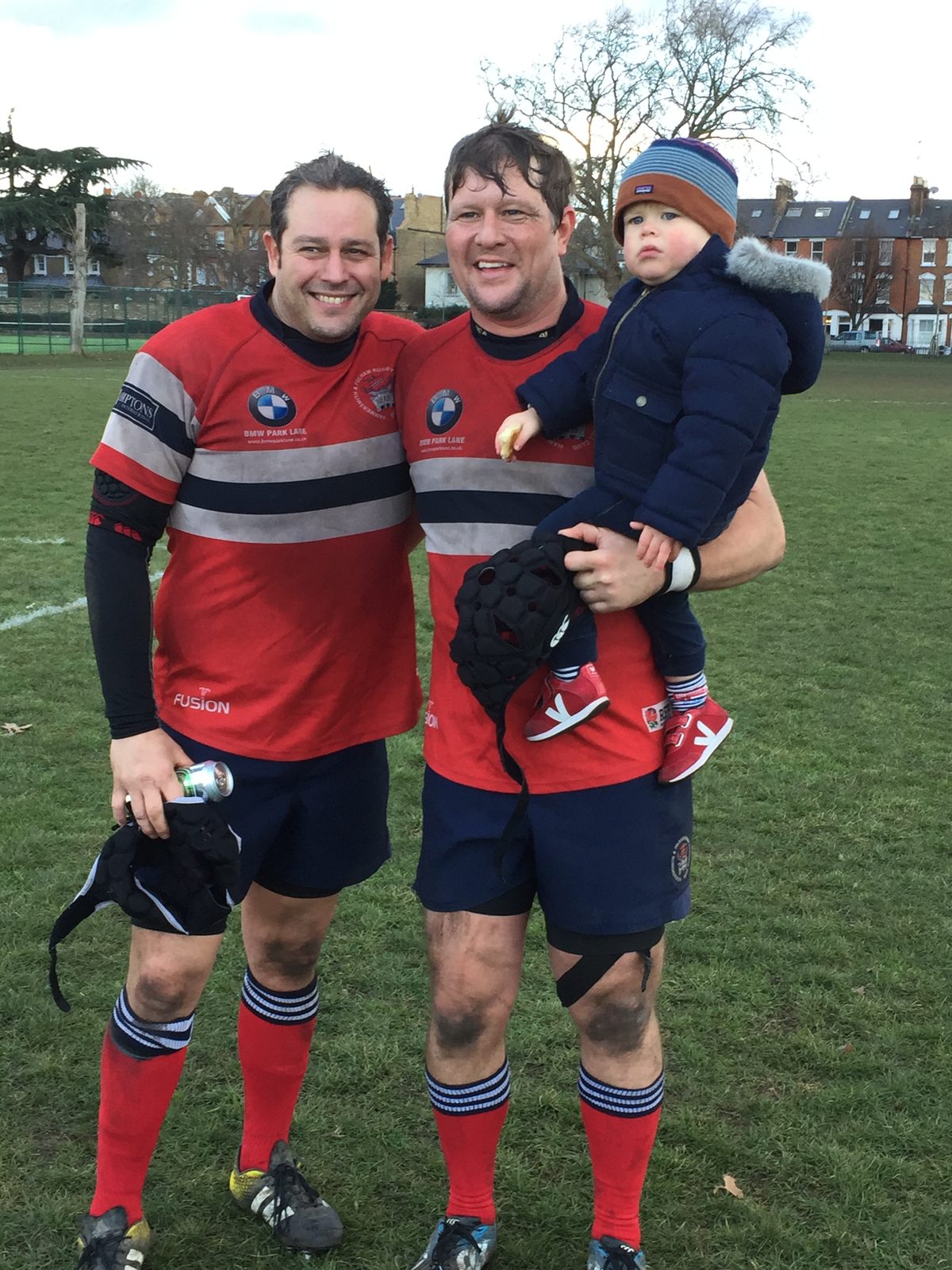 Justin in a muddy rugby kit after a match, holding his young son